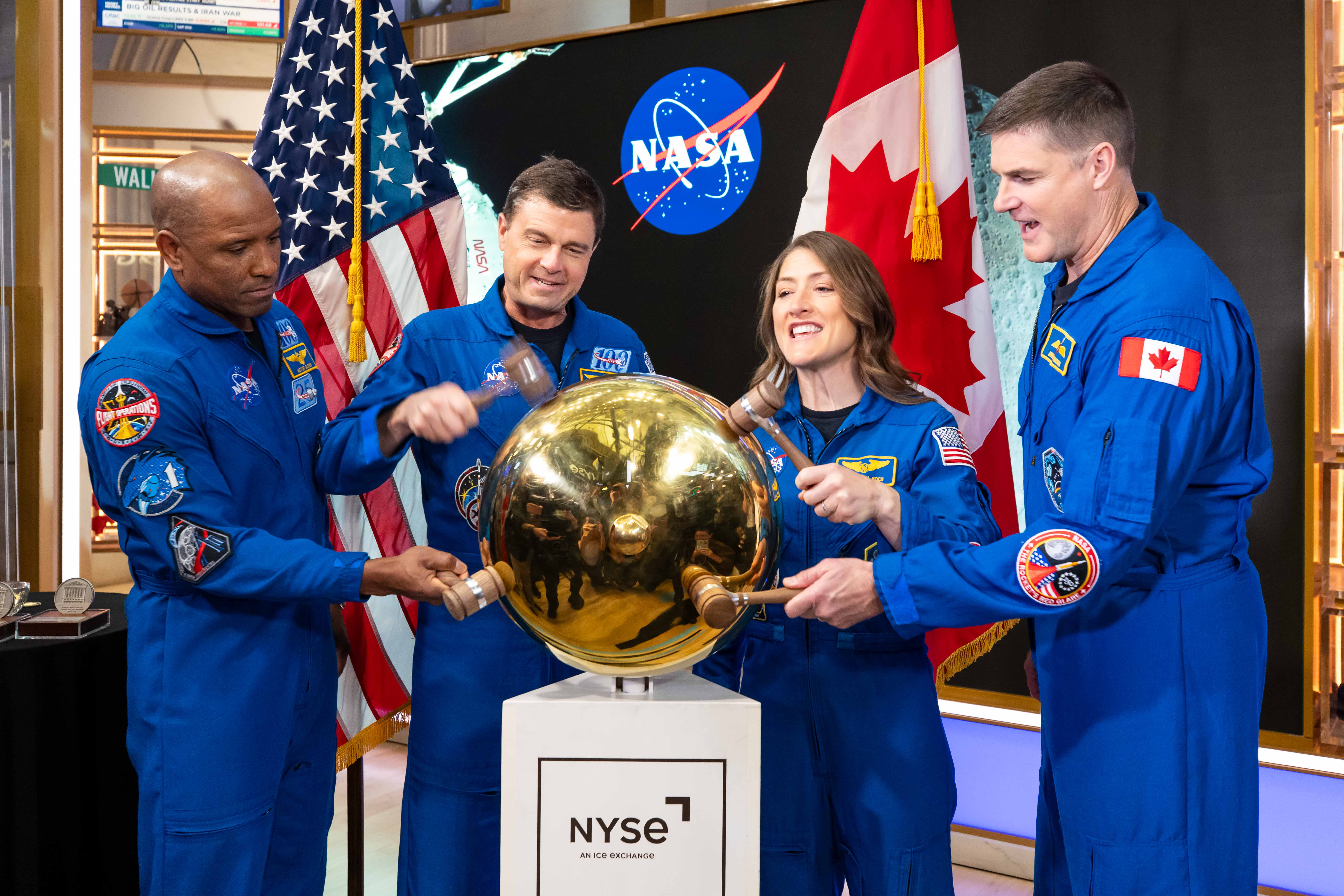 Four astronauts in blue flight suits strike a gold sphere with wooden gavels on an NYSE pedestal, in front of a NASA logo and U.S. and Canadian flags.