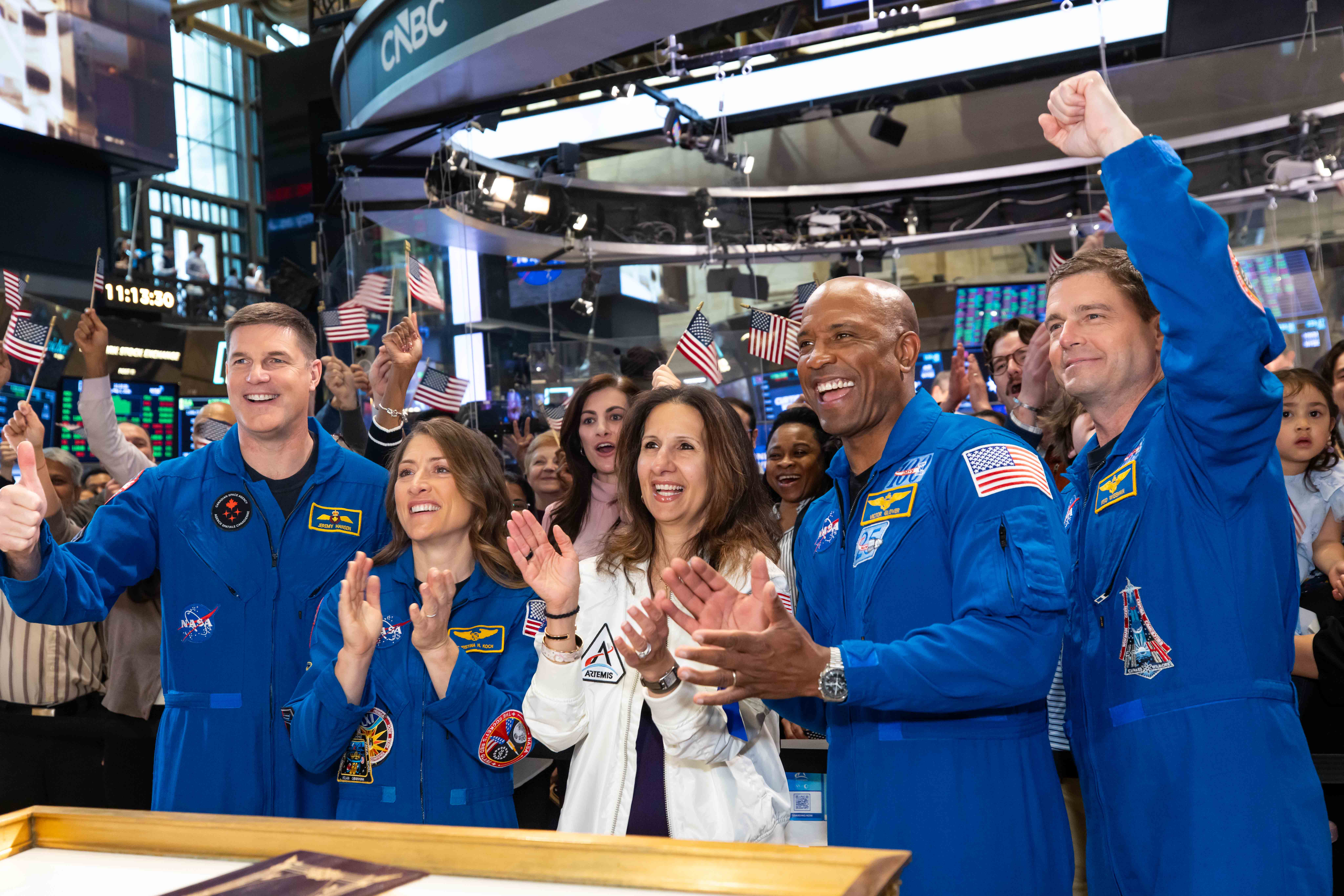 Four astronauts in blue NASA flight suits cheer and applaud with a crowd waving small American flags on the NYSE trading floor, one giving a thumbs-up and another raising a fist.
