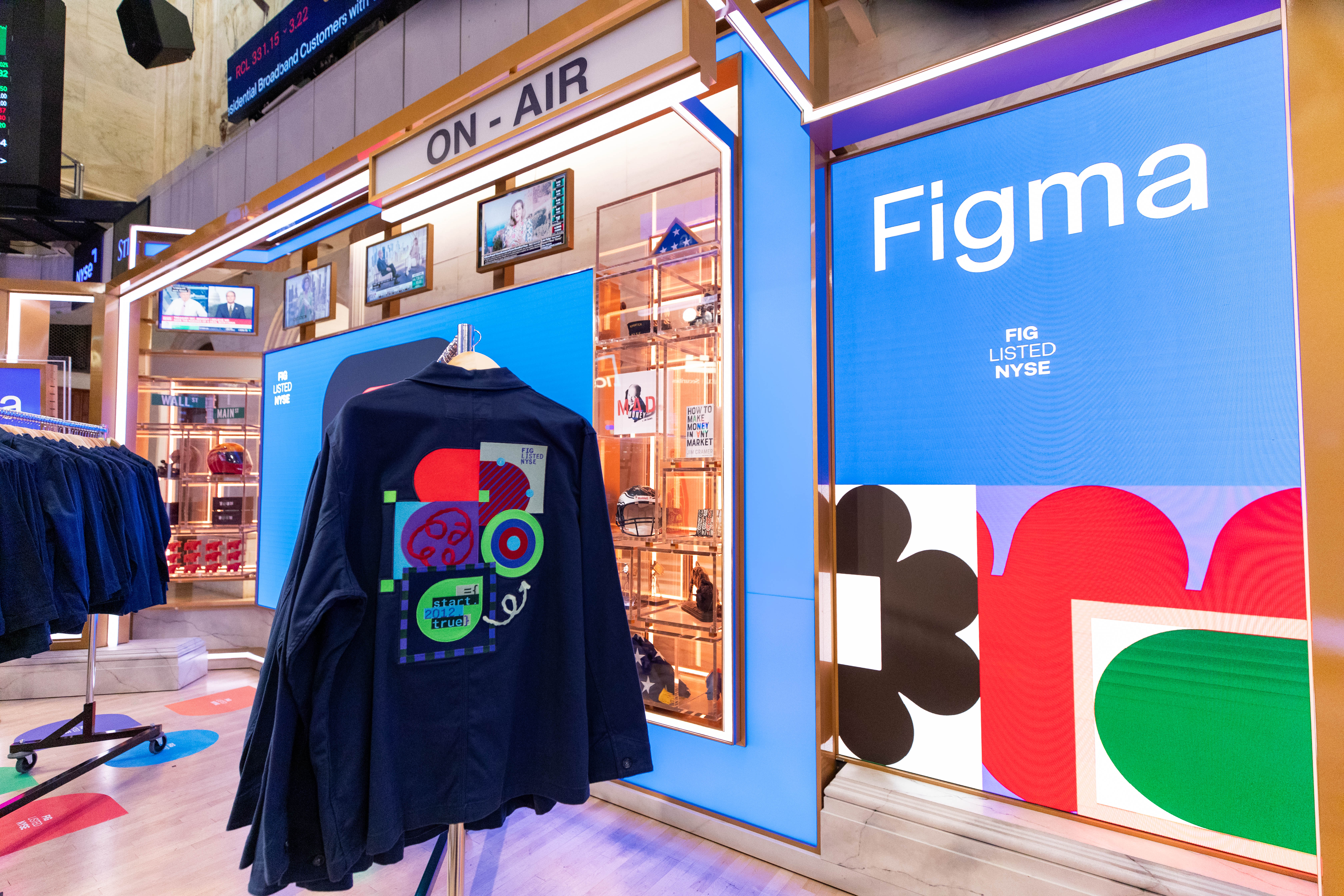 A navy jacket with a colorful embroidered "FIG Listed NYSE" patch hangs on a rack in front of a blue Figma display on the NYSE trading floor.