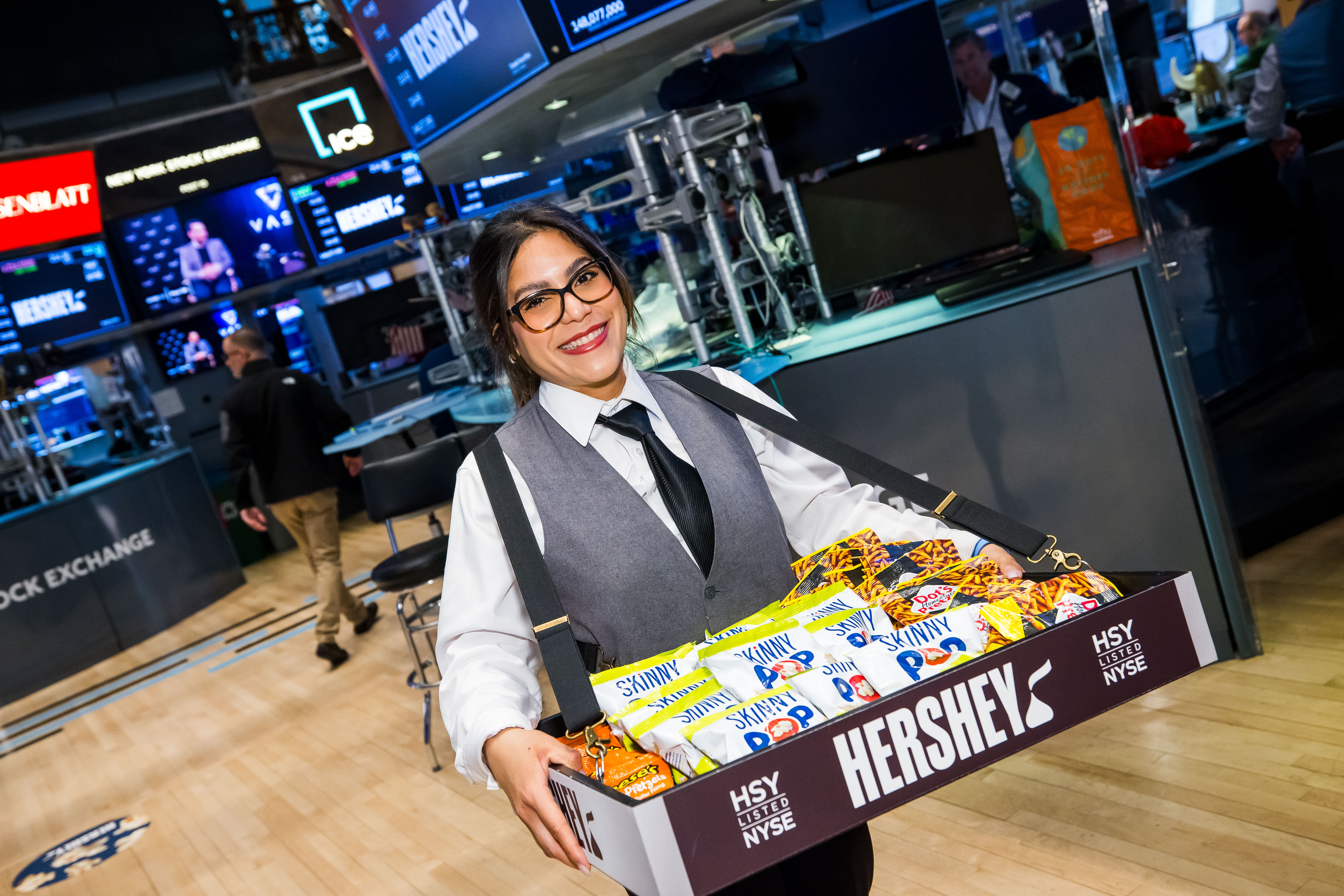 A smiling woman in glasses, a gray vest and a black tie carries a strap-mounted tray of snacks branded "Hershey HSY Listed NYSE" across the New York Stock Exchange trading floor.