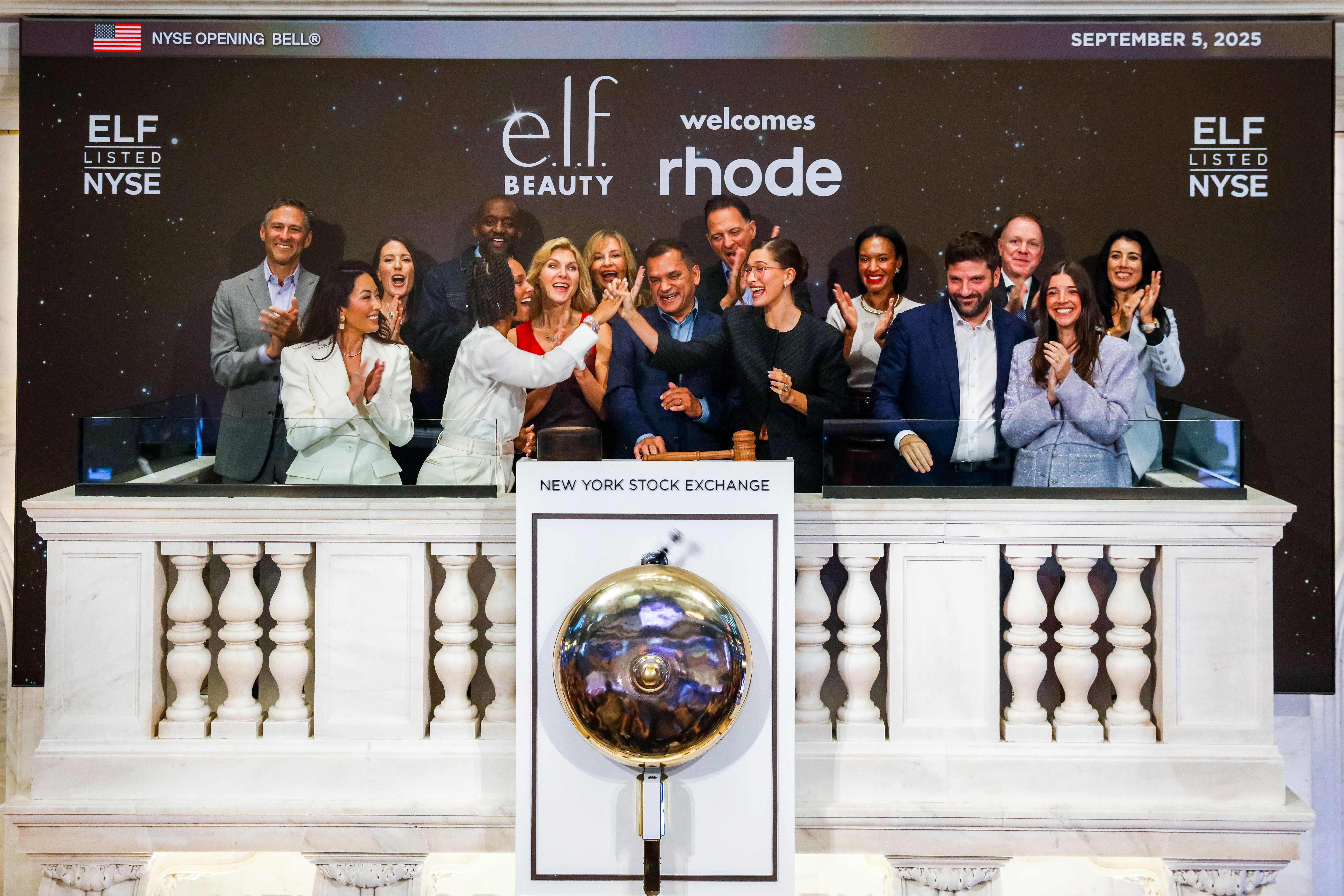 A group of about sixteen people cheer and applaud on the NYSE bell-ringing balcony on September 5, 2025, beneath a backdrop reading "e.l.f. Beauty welcomes rhode," as two women in the center high-five.