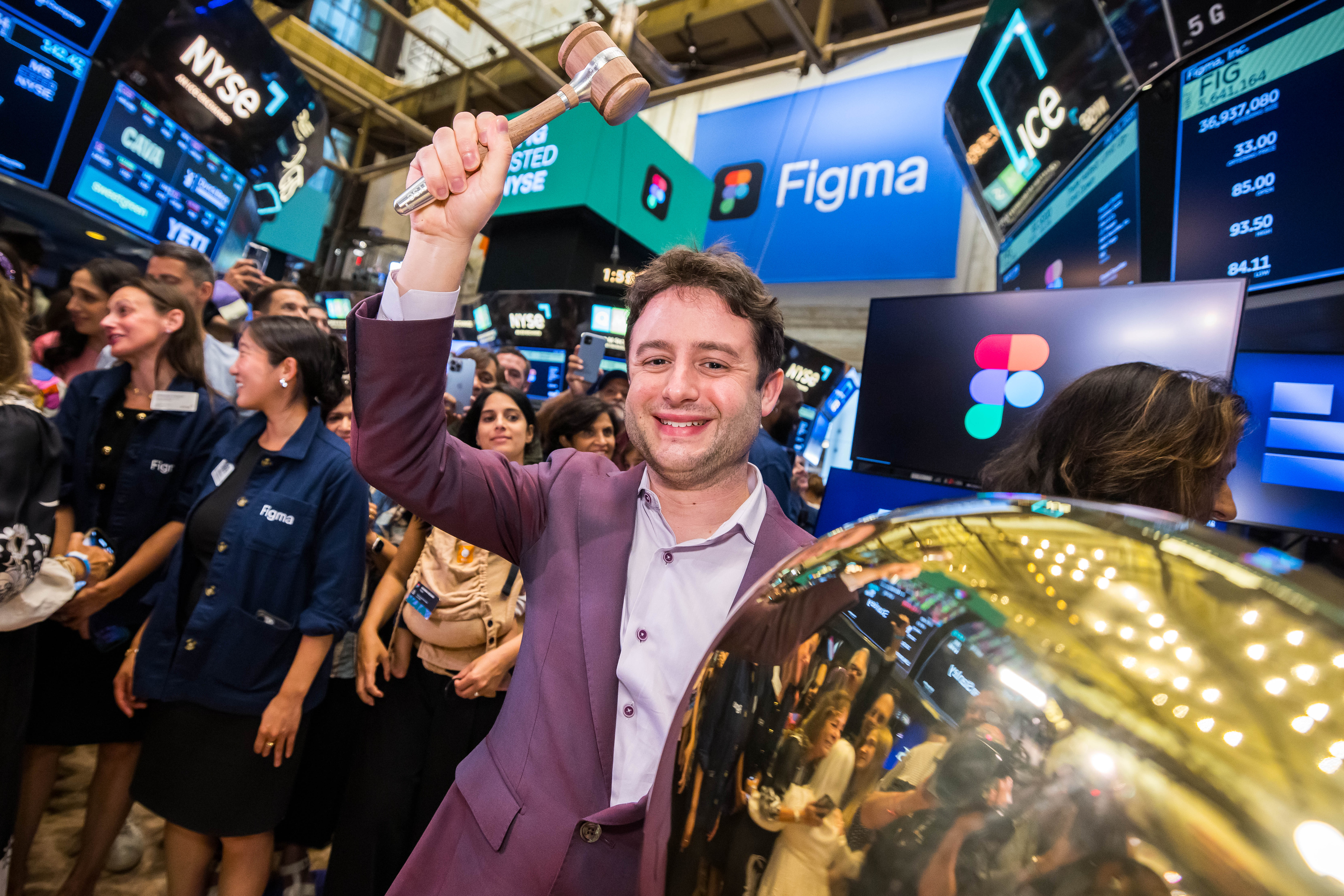A smiling man in a purple blazer raises a wooden gavel high beside a large gold bell on the NYSE trading floor, with Figma-branded screens and a crowd of cheering colleagues behind him.