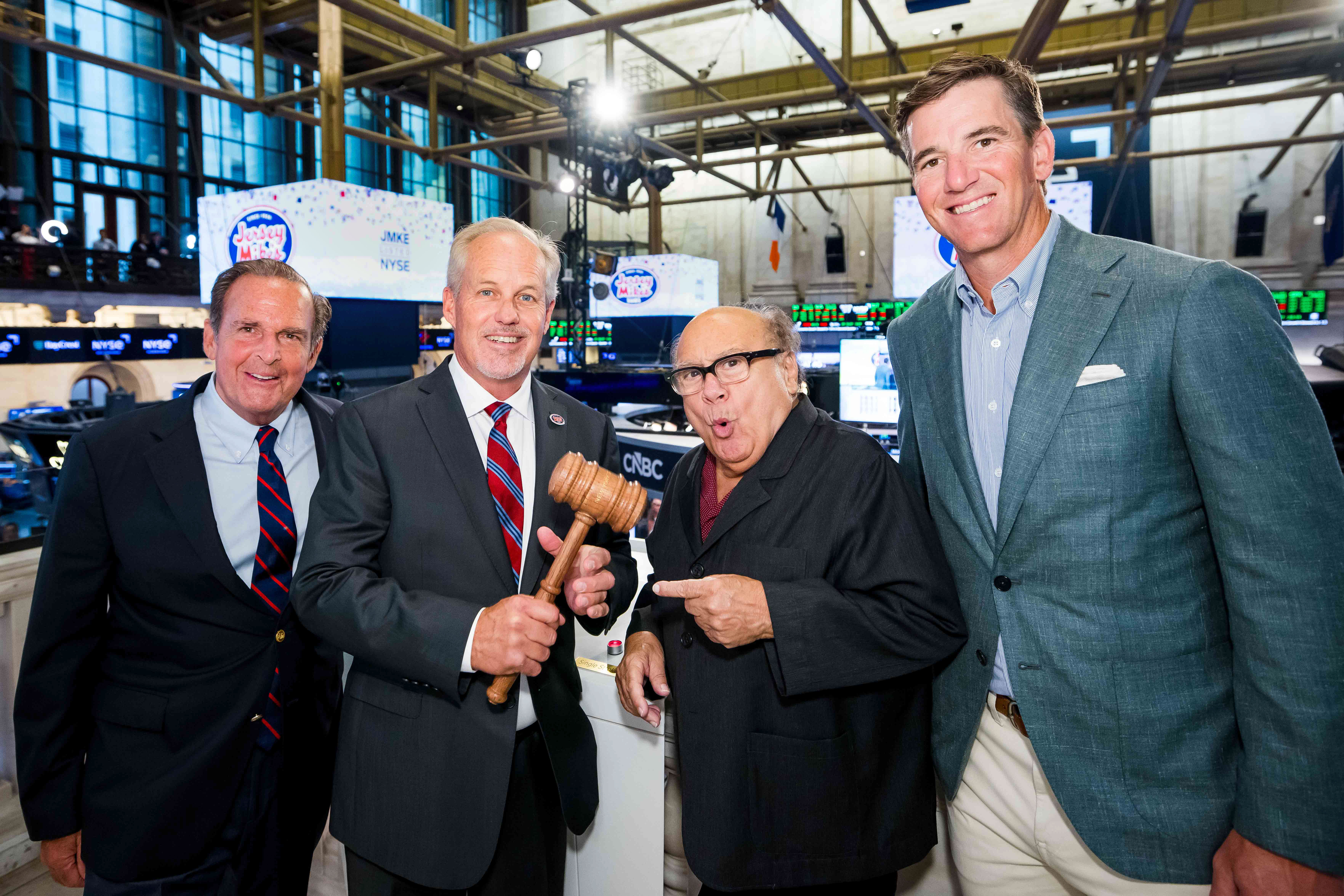 Four men pose together on the NYSE balcony, one holding a wooden gavel while another points at it with a playfully surprised expression, with Jersey Mike's "JMKE Listed NYSE" screens behind them.