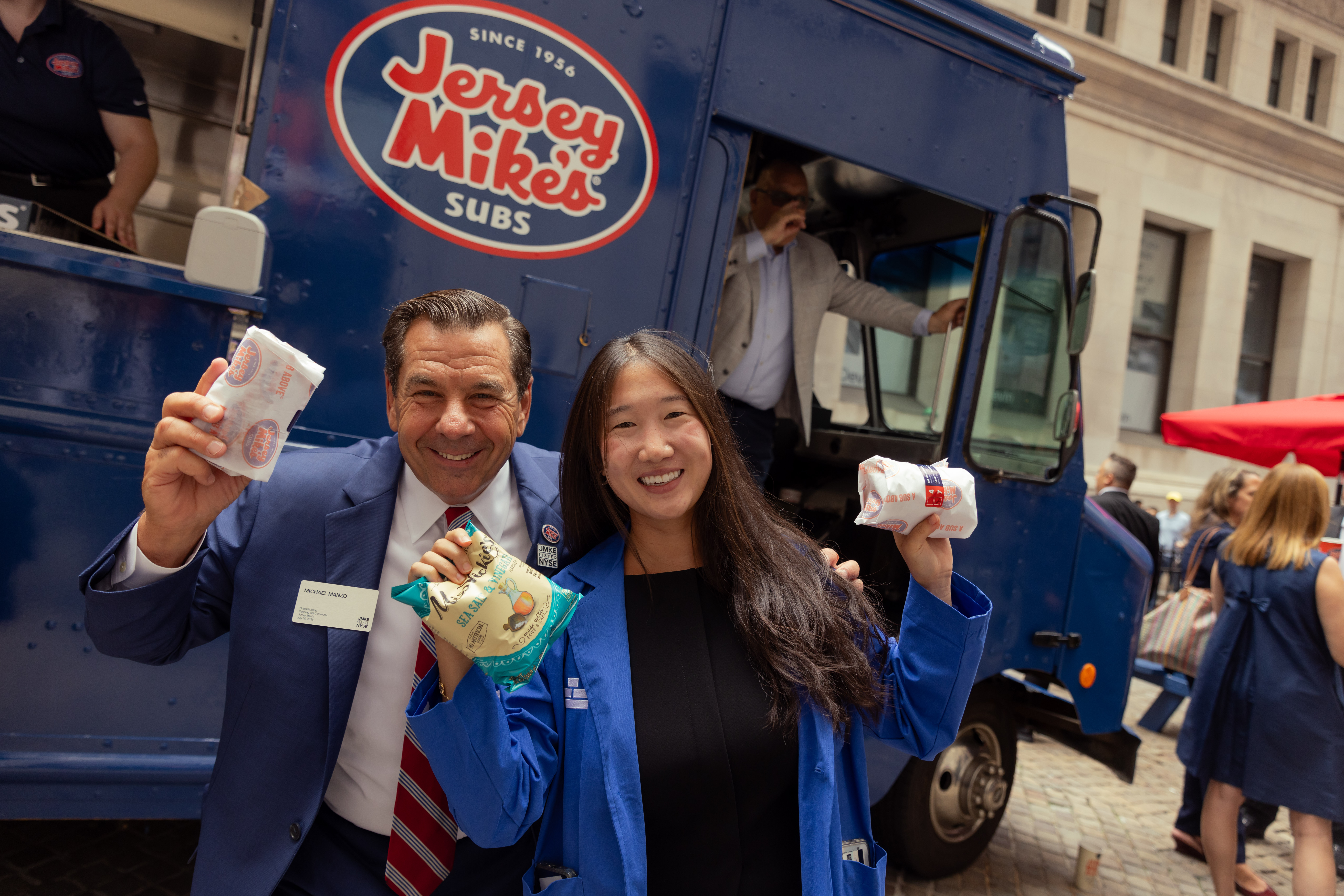 Two smiling people hold up wrapped subs and a bag of chips in front of a blue Jersey Mike's Subs food truck on a cobblestone street.