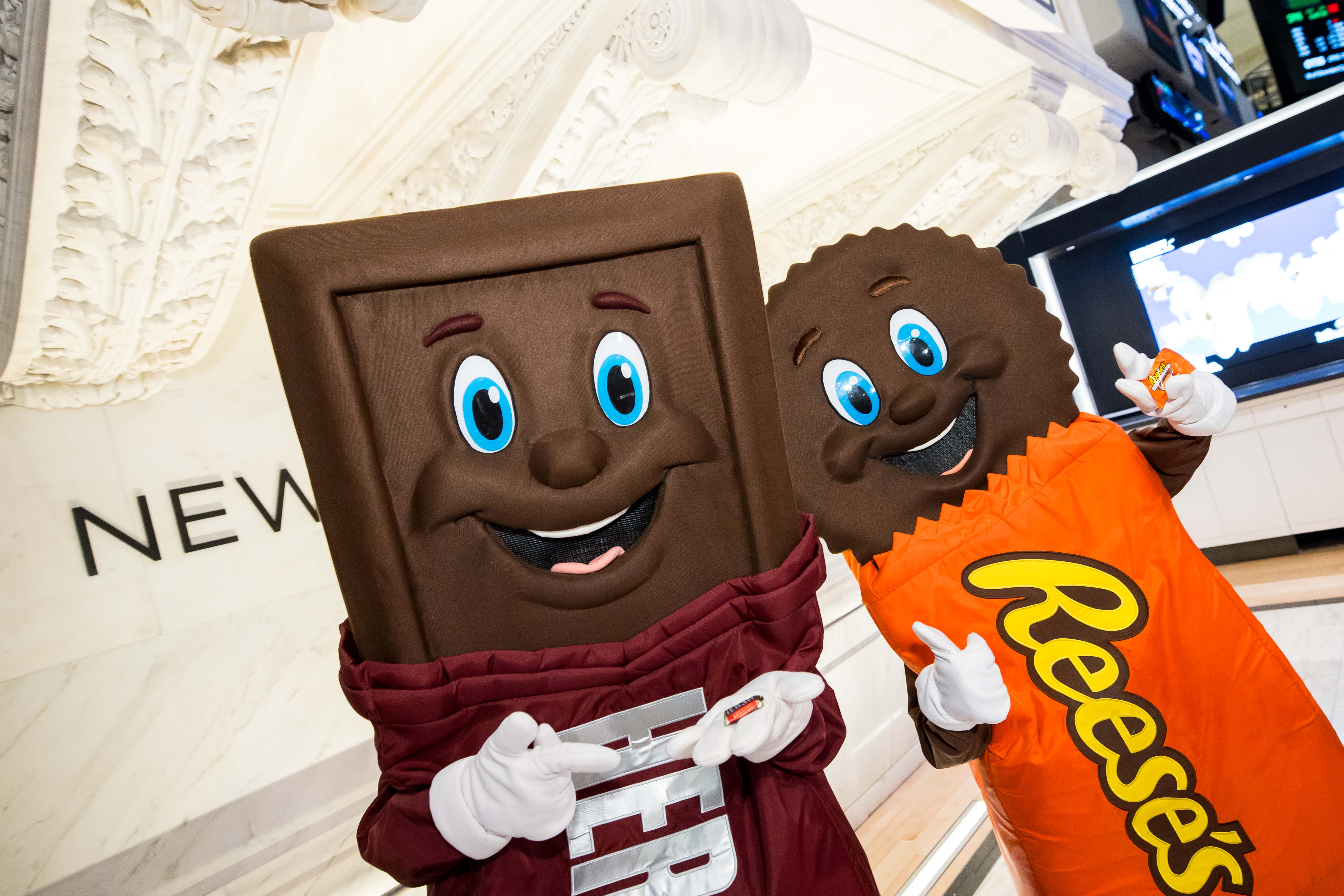 Hershey's chocolate bar and Reese's peanut butter cup mascots posing together inside the New York Stock Exchange, each holding a small piece of candy.