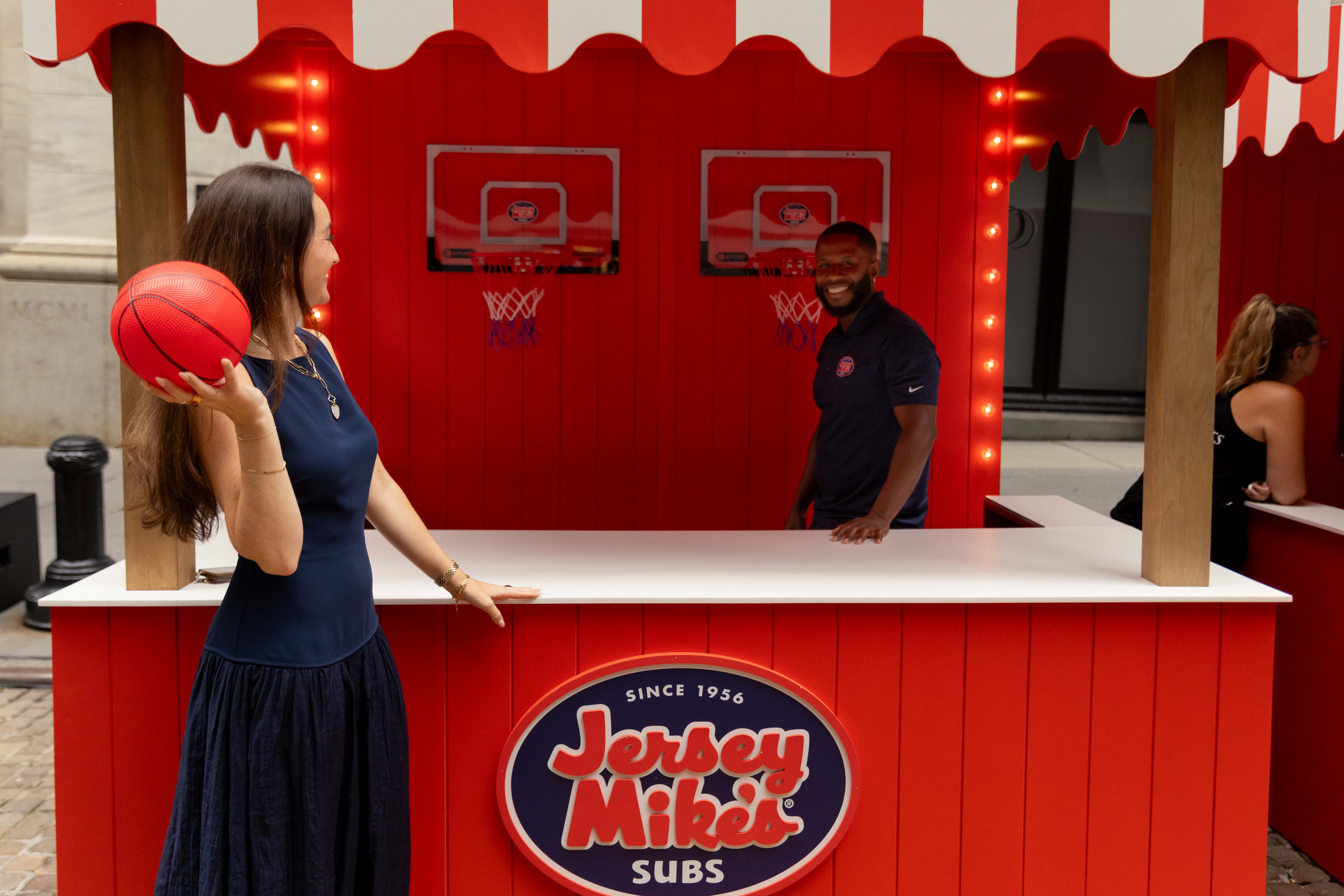 A woman in a navy dress prepares to toss a small red basketball at a hoop game inside a red-and-white Jersey Mike's Subs booth, while a smiling man in a navy polo watches from behind the counter.