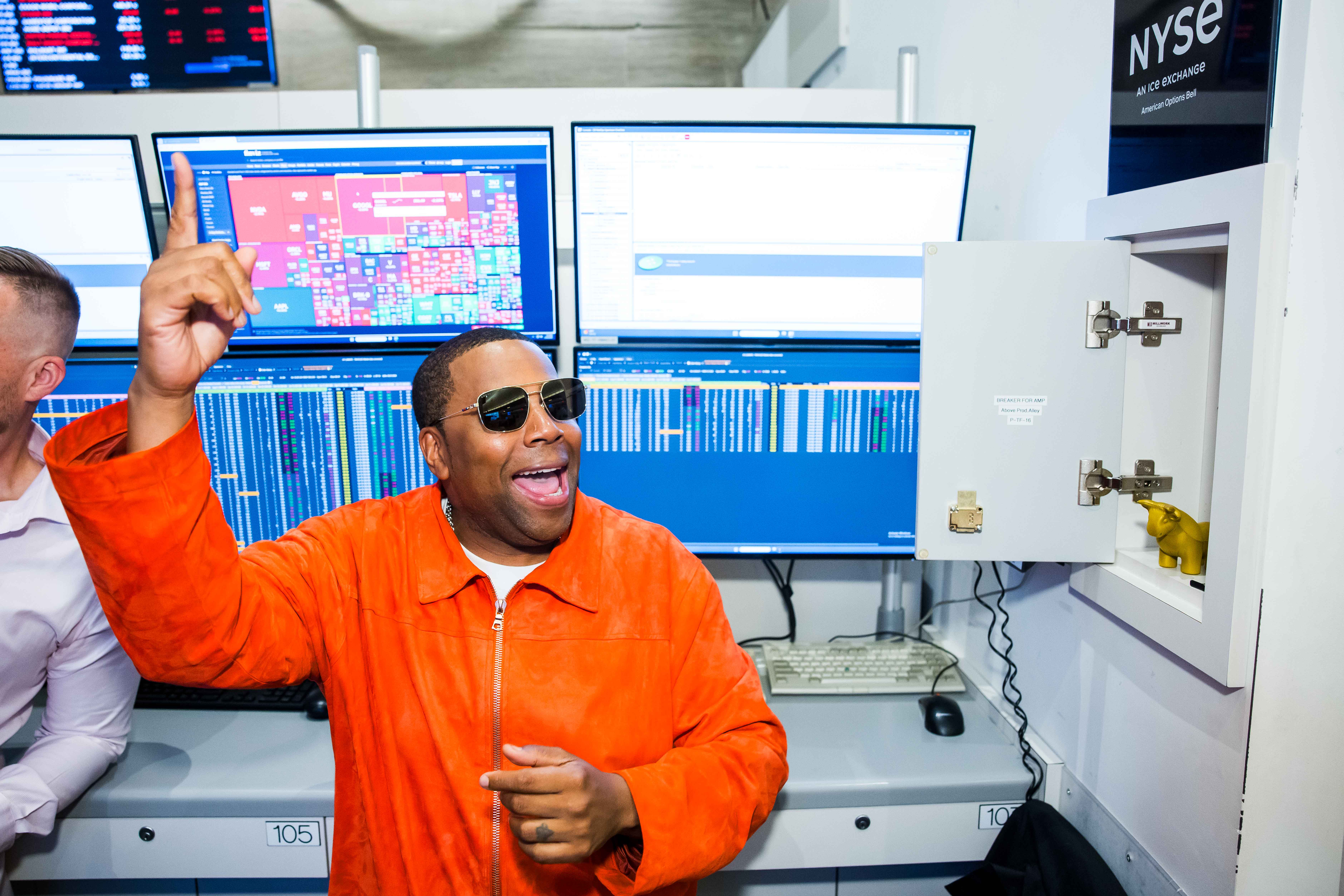 Kenan Thompson, in an orange suede jacket and aviator sunglasses, points one finger skyward and smiles broadly in front of trading monitors at the NYSE American Options trading floor.