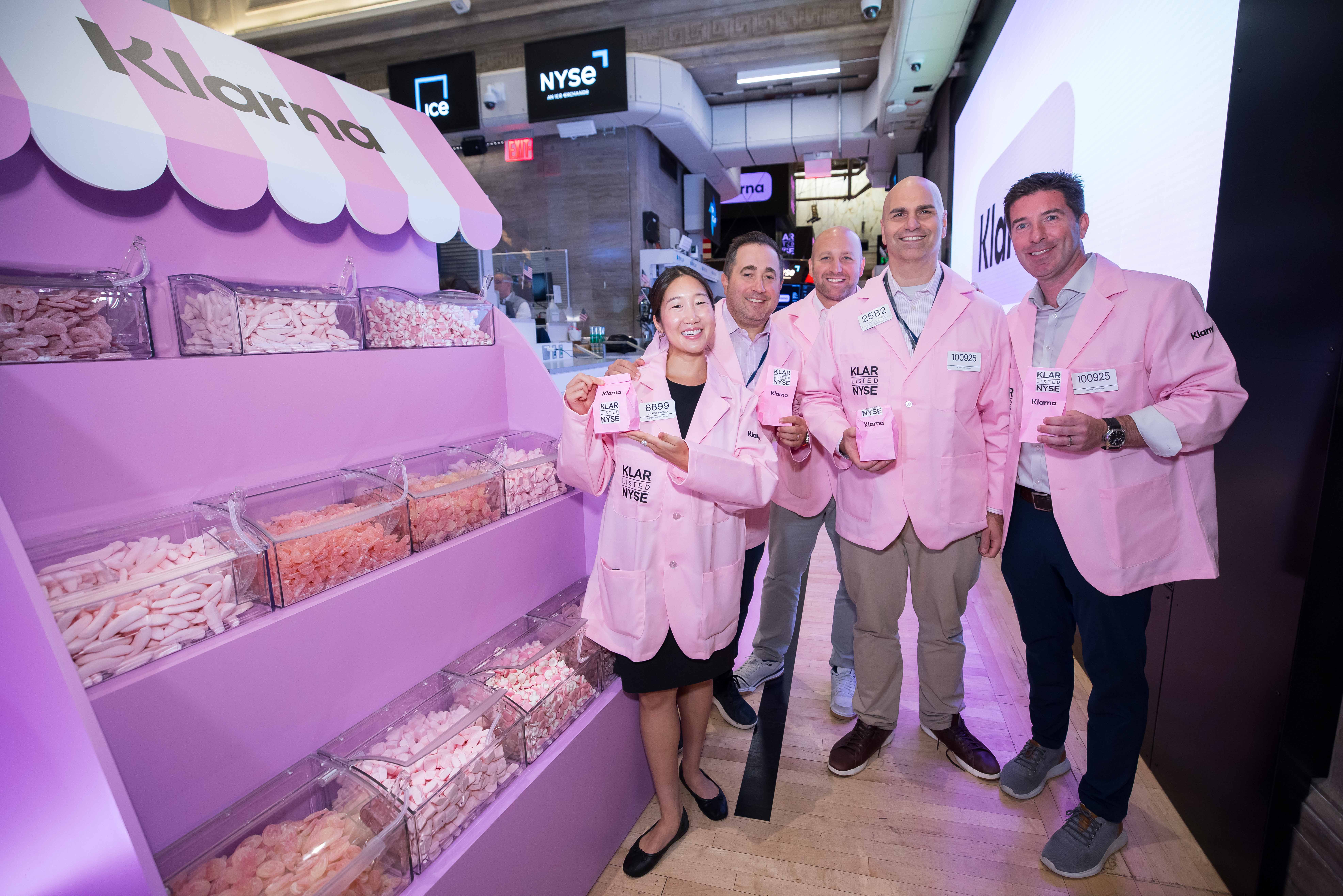 Five smiling people in pink "KLAR Listed NYSE" lab coats hold small pink candy bags beside a pink Klarna candy-shop display on the New York Stock Exchange floor.