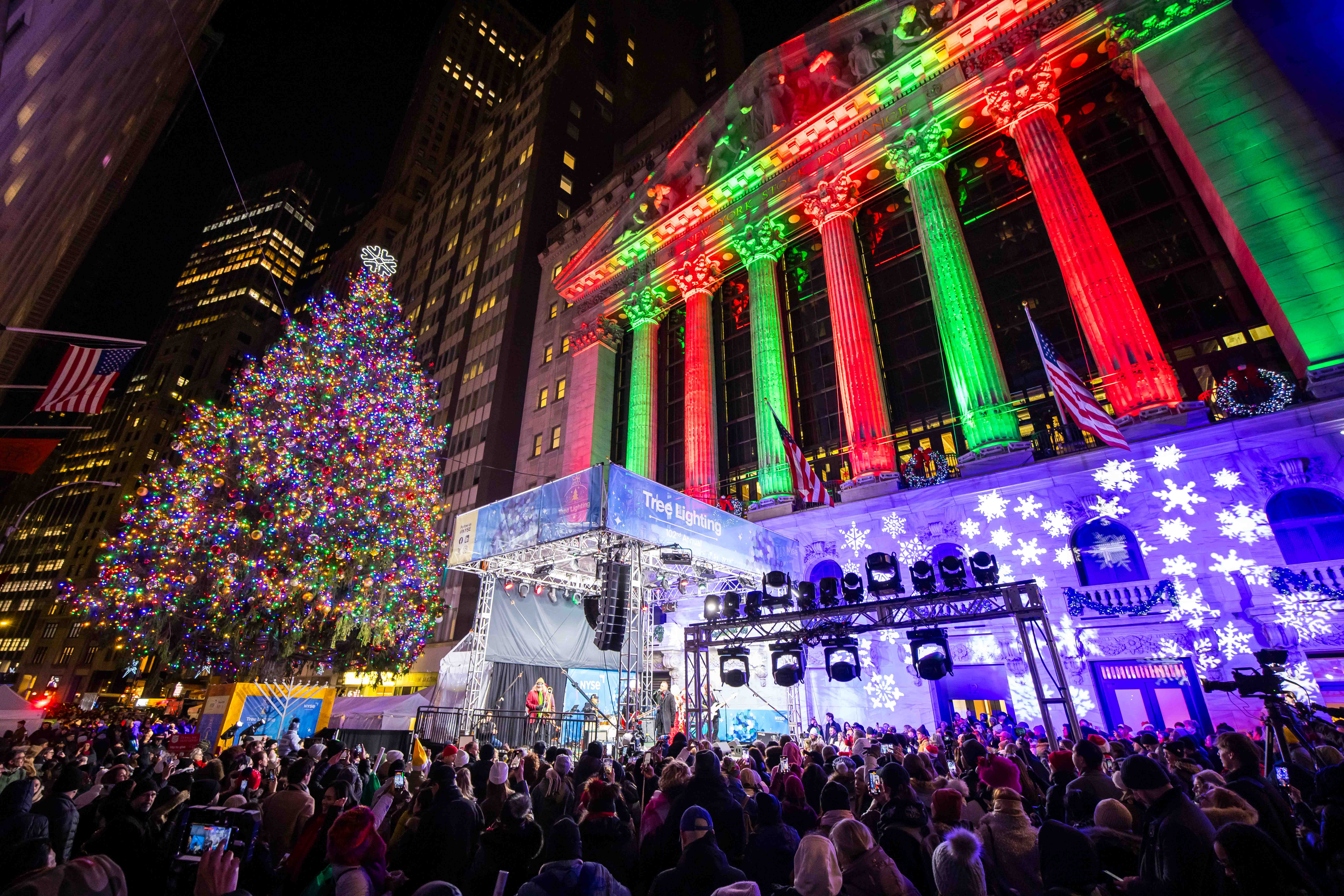 A large crowd gathers at night outside the New York Stock Exchange for a tree lighting, with a huge, brightly lit Christmas tree beside the building's columns glowing red and green.