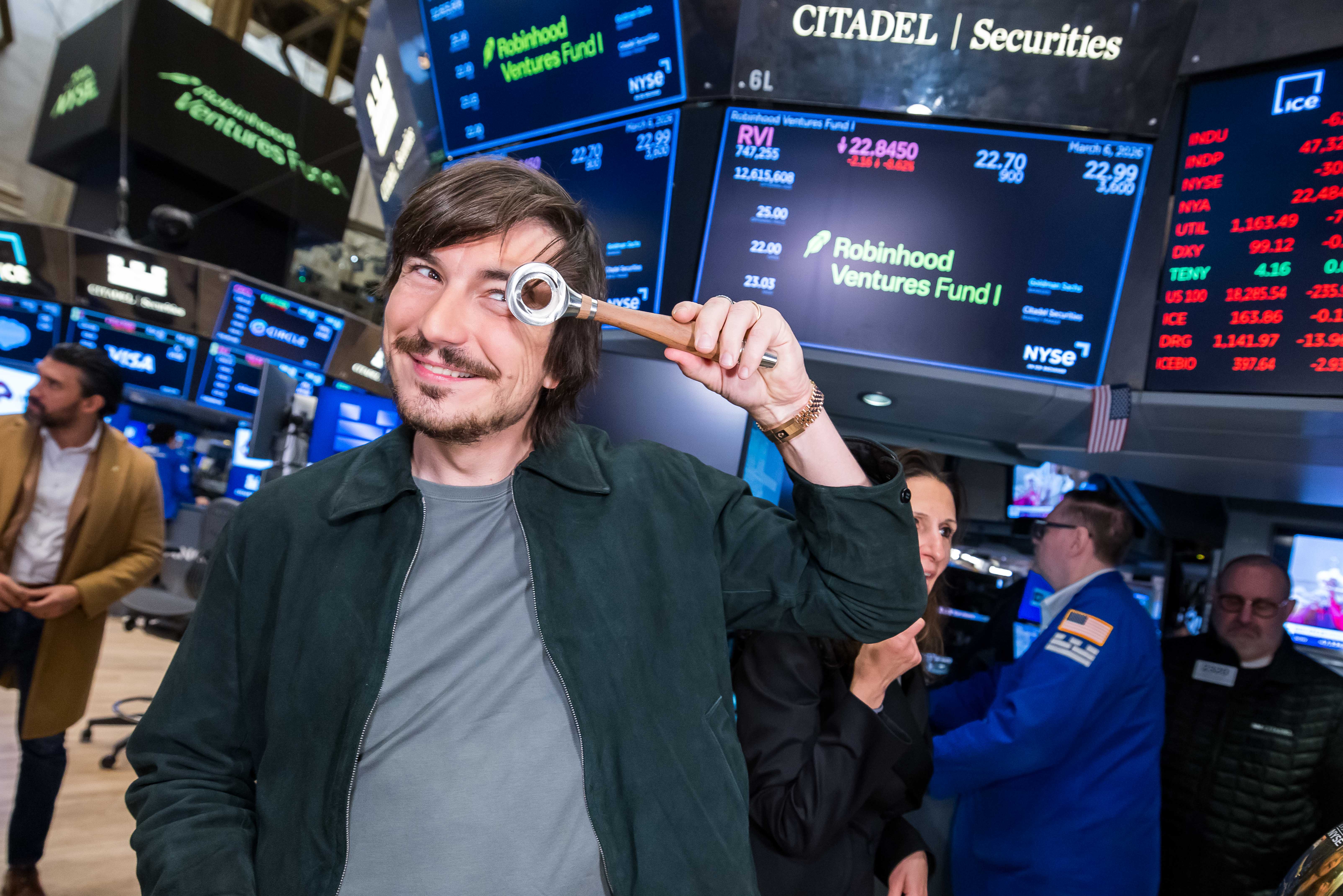 A smiling man in a dark green jacket holds a wooden-handled silver ring up to his eye like a monocle on the NYSE trading floor, in front of screens showing Robinhood Ventures Fund I.
