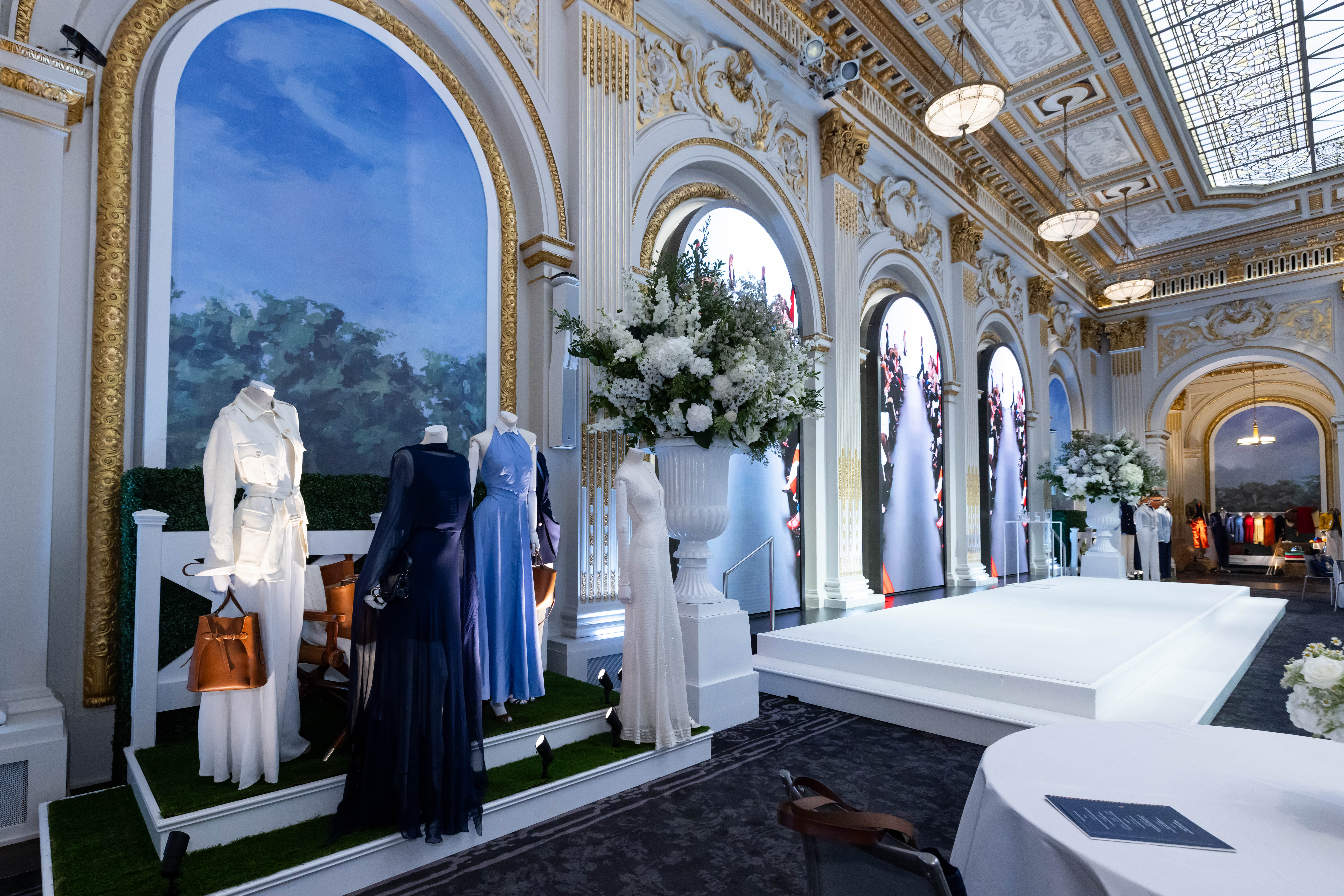 A fashion presentation set up in an ornate ballroom inside the NYSE building, with mannequins in white, navy and light blue garments on a grass-covered platform beside a white runway.