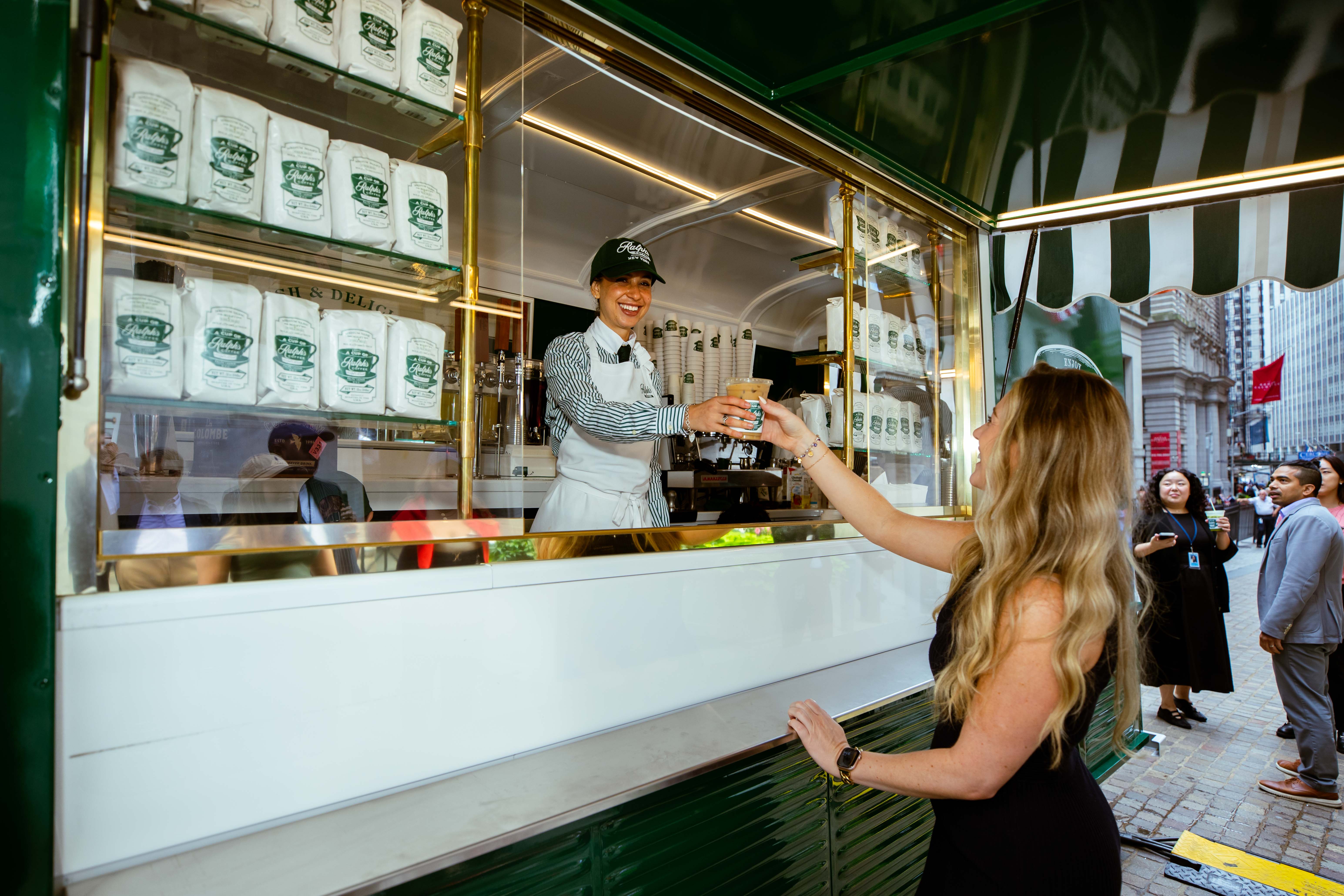 A smiling barista in a Ralph's Coffee cap and white apron hands an iced coffee to a woman through the window of a green-and-white coffee truck on a city street.