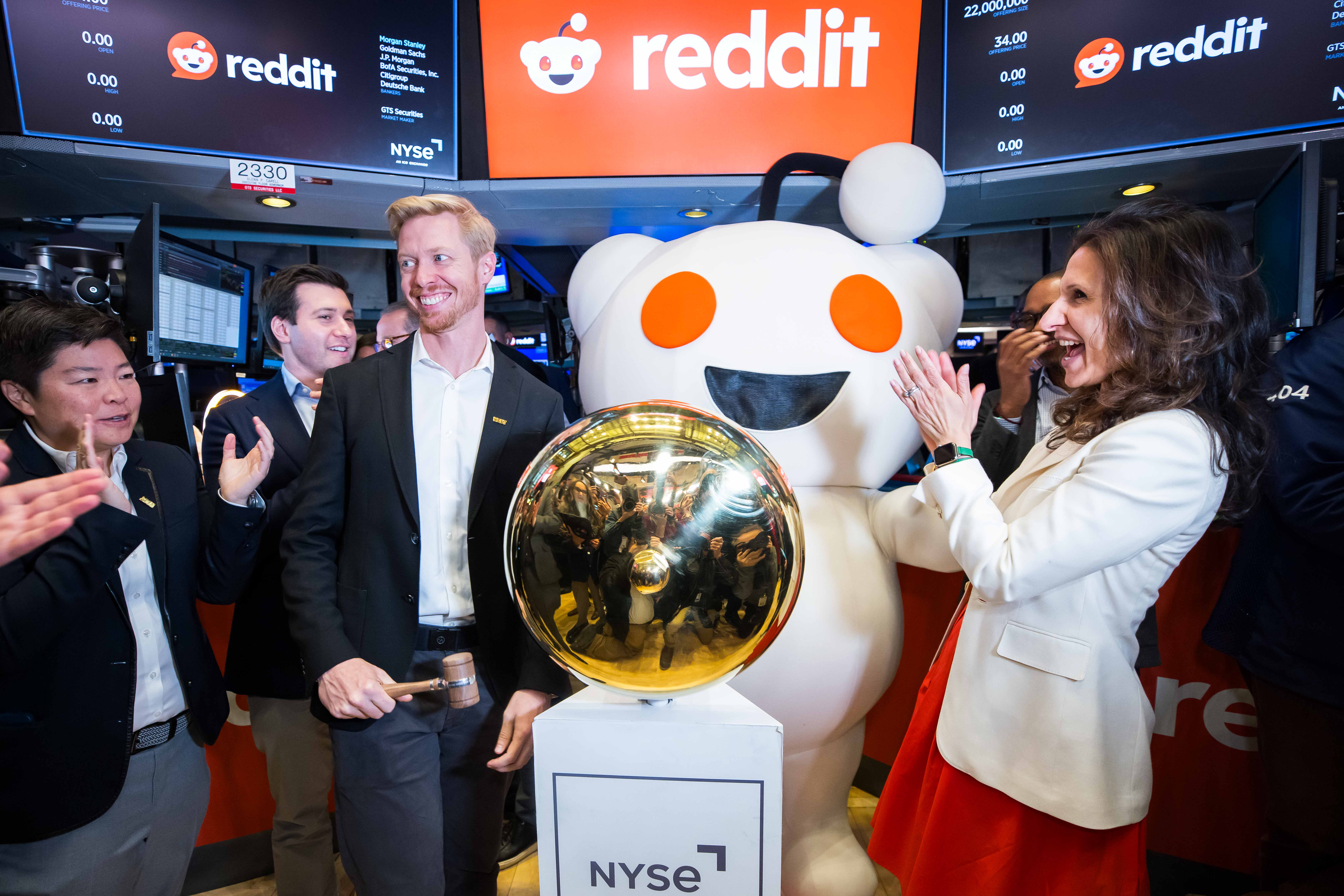 A man holding a wooden gavel smiles beside a gold NYSE bell as colleagues and Reddit's Snoo mascot applaud on the trading floor, beneath screens showing the Reddit logo.
