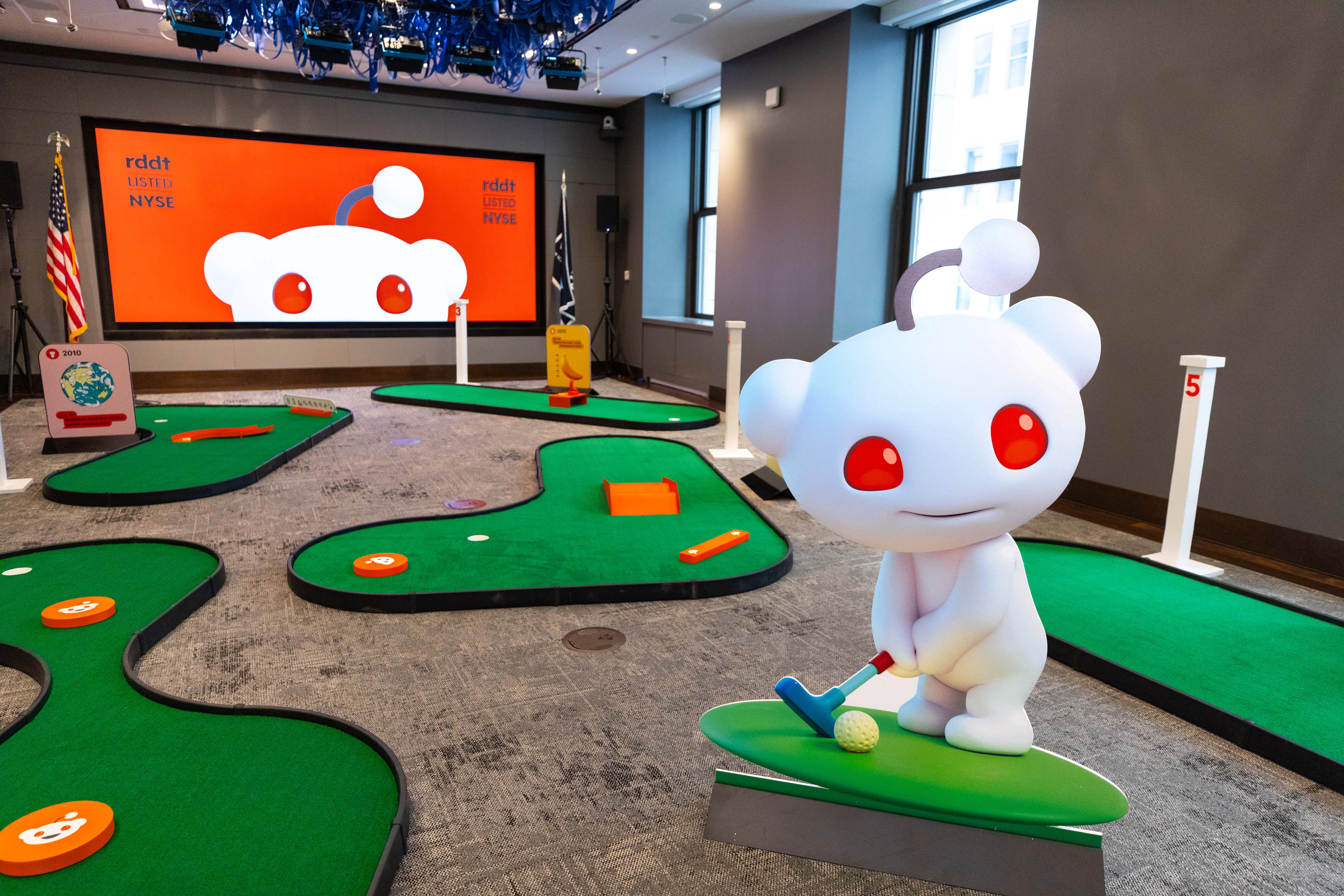 A life-size Reddit mascot, Snoo, stands in a putting pose beside a mini golf course set up in an event room, with a large orange screen reading "rddt Listed NYSE" behind it.
