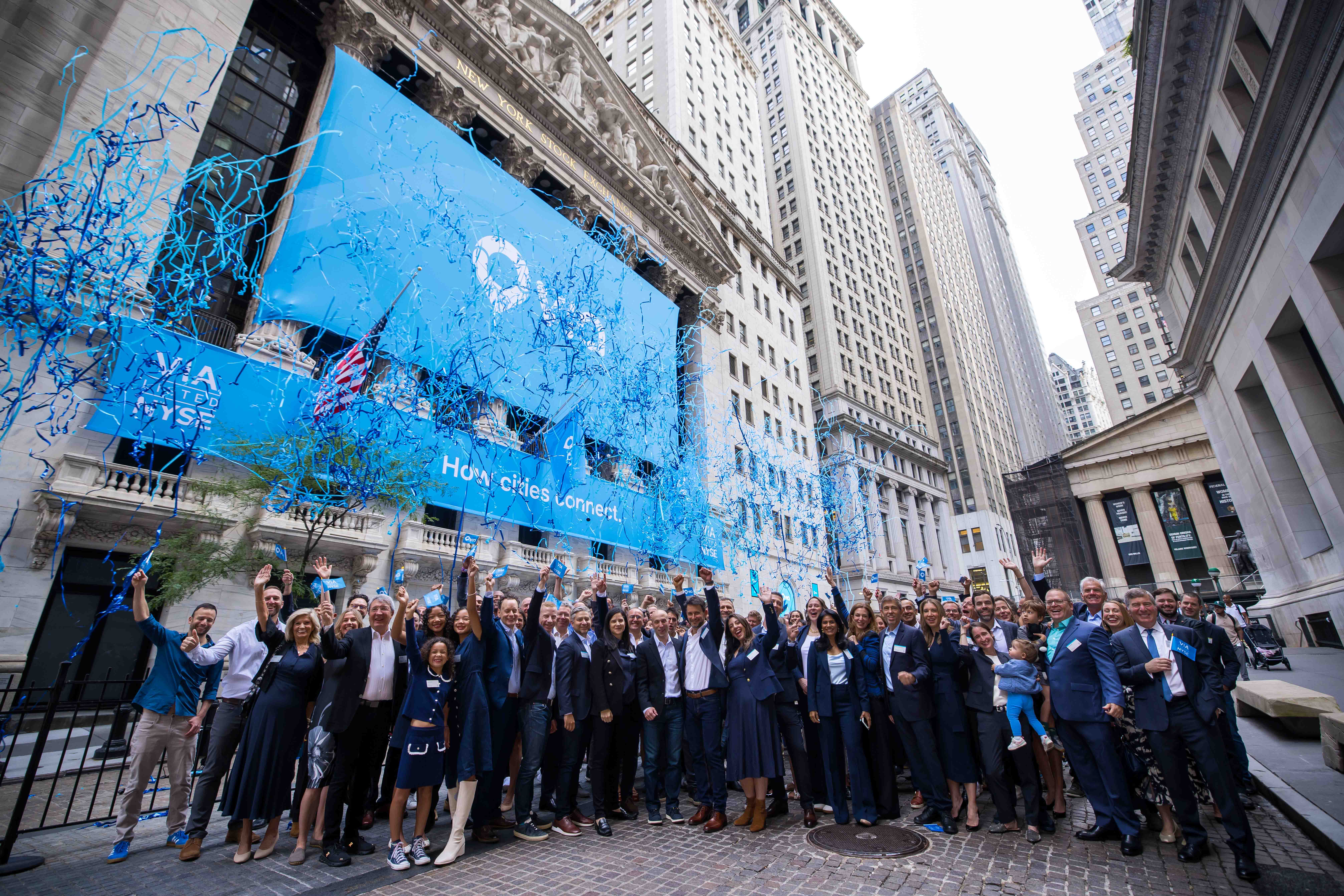 A large crowd of people in business attire cheer and raise their arms on Wall Street as blue streamers fly in front of the New York Stock Exchange, which is draped with blue Via banners reading "How cities connect."